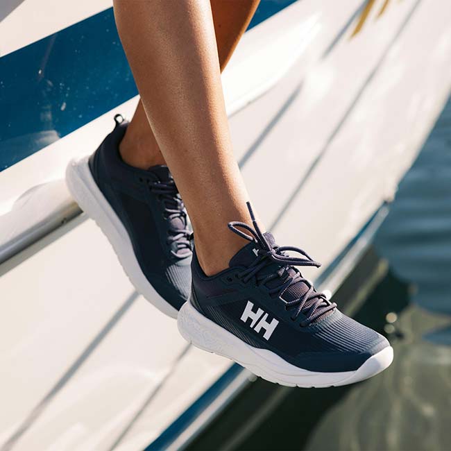 A close-up of a person's legs wearing Helly Hansen athletic sneakers on a boat. The shoes are dark navy blue with a white sole and a white 'HH' logo on the side.